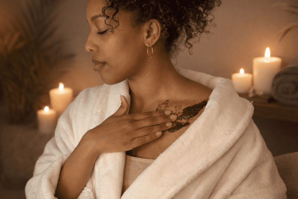 A woman wrapped up in clean fresh robes applying moroccan body scrub product onto her chest skin while surrounded by aromatic candles in a relaxing spa environment